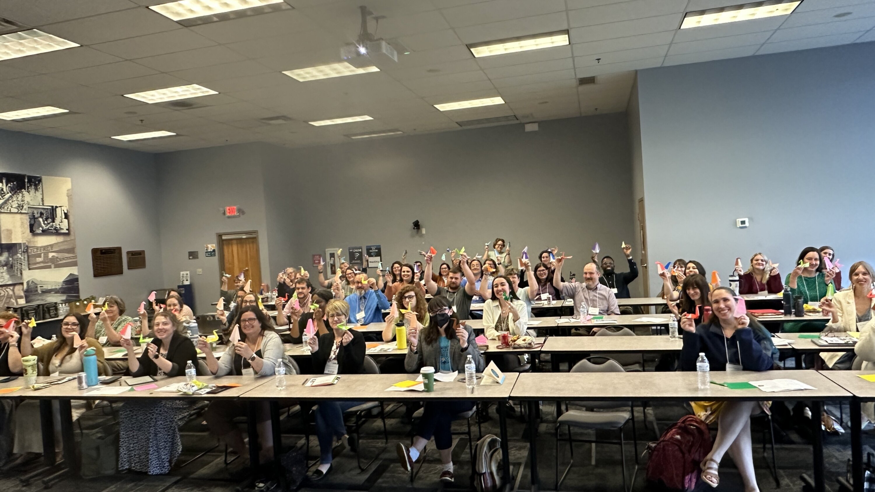 A large group of MakerFest attendees seated at tables and smiling while holding up colorful origami creations made during a hands-on afternoon session at the State Library of Ohio.