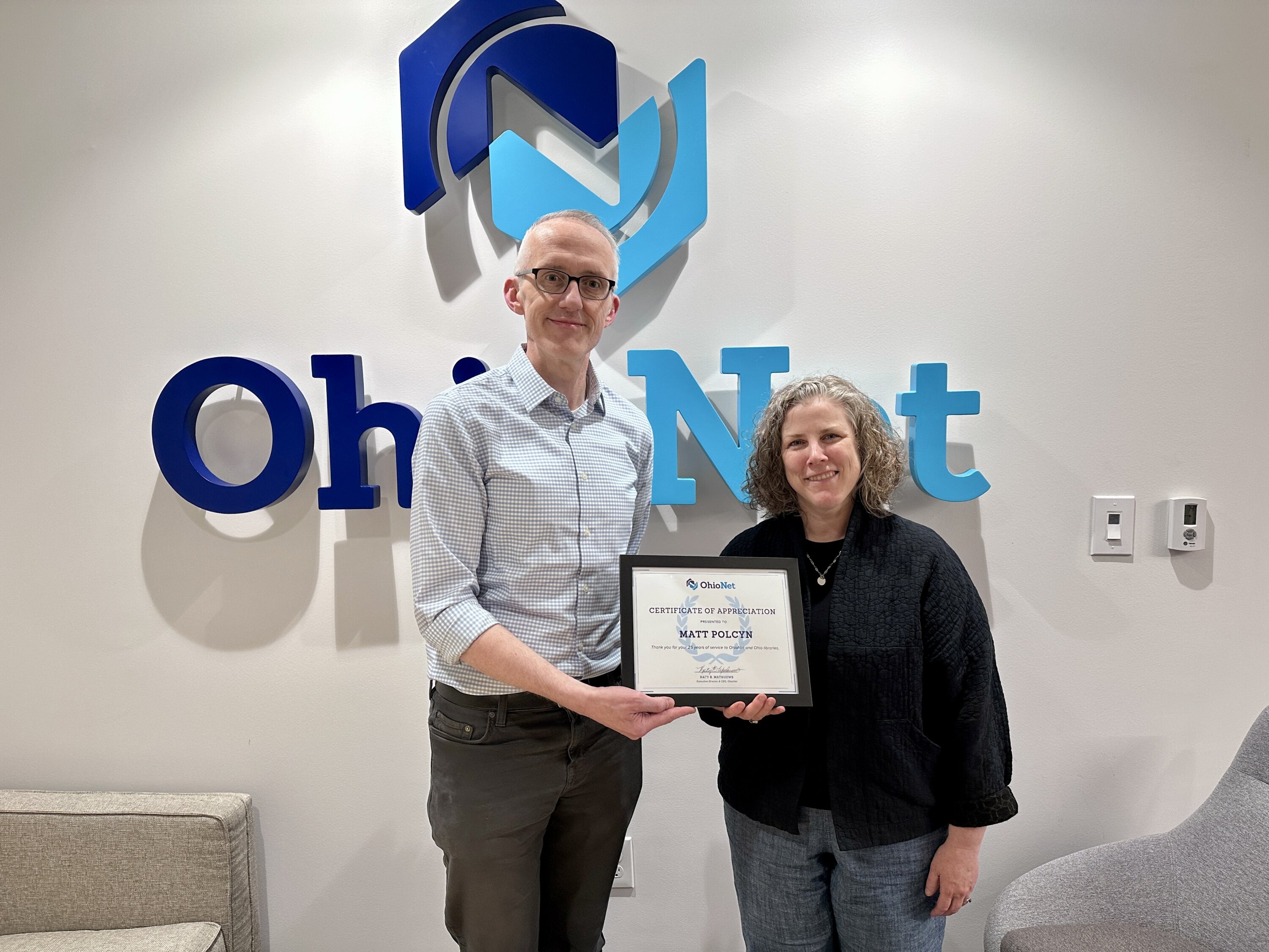 Matt Polcyn, OhioNet Director of Technology Services, accepts a certificate for 25 years of service from Katy Mathuews, OhioNet Executive Director & CEO. They are standing and smiling in front of a white wall with OhioNet signage behind them.